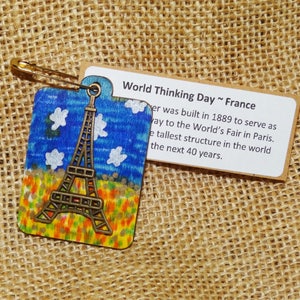 May include: A wooden Eiffel Tower ornament with a blue sky and white clouds painted background. The ornament is attached to a gold safety pin. The text "World Thinking Day ~ France" is printed on a brown card.