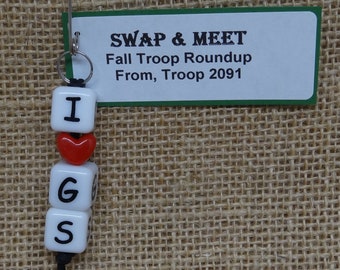 May include: A white keychain with the letters "I", "G", and "S" spelled out on individual beads. A red heart bead is between the "I" and "G" beads. A green tag with the text "SWAP & MEET Fall Troop Roundup From, Troop 2091" is attached to the keychain.