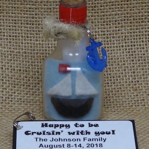 May include: A small glass bottle with a cork stopper and a blue anchor charm. The bottle contains a white sailboat with a red flag. The bottle is tied with twine and has a black tag with the text "Happy to be Cruisin' with you! The Johnson Family August 8-14, 2018".