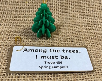 May include: A green plastic Christmas tree ornament with a gold wire hanger. The ornament has a tag attached to it that reads "Among the trees, I must be. Troop 456 Spring Campout".