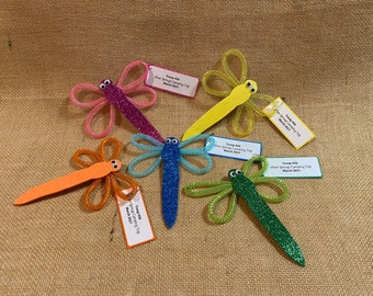 May include: Five colorful dragonfly crafts with glitter and googly eyes. Each dragonfly has a tag attached with the text "Troop 416 Silver Springs Camping Trip March 2021".