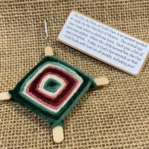 May include: A green, red, and white yarn Ojo de Dios, a traditional Mexican craft, is made with wooden sticks and yarn. The text on the card reads: "The Ojo de Dios, or God's Eye, is an ancient symbol made by the Huichol of Mexico. The central eye was made when a child was born. Each year, a bit of yarn was added. When the child turned five, the Ojo was complete. Today, a God's Eye reminds us that God looks with love on all people, including you!"