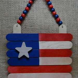 May include: A wooden American flag made from popsicle sticks, painted red, white, and blue, with a white star in the blue section. The flag is hanging from a string of red and blue beads.