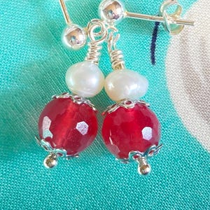 May include: A pair of dangle earrings featuring a red faceted bead, a white pearl, and a silver-colored metal accent. The earrings have a post-back closure. The red beads have a faceted surface.