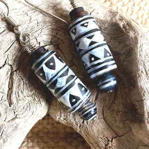 May include: A pair of handmade earrings featuring cylindrical beads with a black and white geometric pattern. The earrings have silver-colored hooks and small, dark wooden accents. The beads are a light blue color.