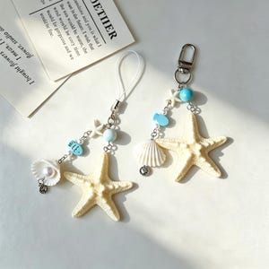 May include: Two beach-themed charms. One has a white seashell with a pearl, a blue fish, and a starfish. The other has a starfish, a blue bead, and a fish. Both have silver-colored hardware.