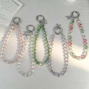 May include: Five beaded keychain straps in various colors: pink, green, clear, and multi-colored. Each strap features a silver-tone ring and a small charm. The beads are square-shaped and strung together to create a decorative accessory.