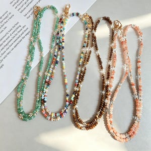 May include: Five beaded necklaces in various colours, including green, multicoloured, brown, and pink. Each necklace features small, colourful beads strung together with gold-coloured clasps. The necklaces are displayed on a light-coloured surface.