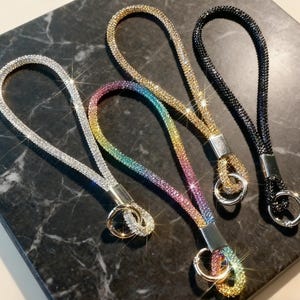 Puede incluir: Cuatro cordones de muñeca brillantes en plata, arcoíris, oro y negro. Cada cordón está cubierto de pequeños pedrería brillantes y presenta un cierre de metal y un llavero. Los cordones se muestran sobre una superficie oscura y veteada.
