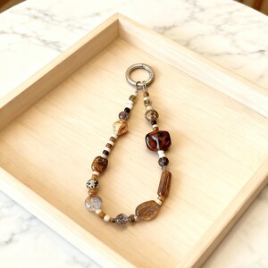 May include: A beaded keyring with a silver clasp. The beads are a mix of brown, tan, and clear, with some speckled and patterned. The keyring is displayed on a light wooden tray.