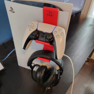 Peut inclure: Une console PlayStation 5 blanche et noire avec un support de manette rouge. Une manette blanche et noire et un casque noir sont fixés au support. La PlayStation 5 a une base noire et une coque blanche.