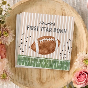 May include: A square greeting card featuring a beige and white striped background with the text Donald's FIRST YEAR DOWN in dark gray. A brown football illustration sits above a green football field graphic with white yard line markings.