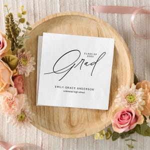 May include: White graduation napkins with the word "Grad" in elegant black script. The napkins also include the text "Class of 20XX", the graduate's name, and the high school name. The napkins are arranged on a wooden tray, surrounded by pink and peach flowers.