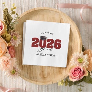 May include: White square napkins with graduation theme. The napkins feature the text "CLASS OF 2026 grad ALEXANDRA" in burgundy and black. The napkins are surrounded by flowers and placed on a wooden tray.