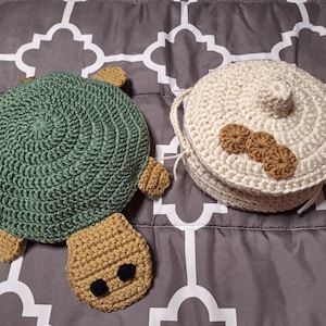 May include: A green and tan crocheted turtle-shaped flat item alongside a round, cream-colored crocheted lidded container with three small tan circular accents on the lid. Both items feature a textured yarn construction with visible stitching.