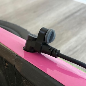 Könnte beinhalten: Nahaufnahme eines schwarzen Netzkabels und -steckers, der an ein Gerät mit einem rosa Streifen angeschlossen ist. Der Stecker hat eine graue Gummiabdeckung. Das Gerät ist schwarz und hat ein Rad.