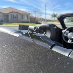 Puede incluir: Una cámara de salpicadero negra montada en el interior de un coche descapotable. La cámara es pequeña y cilíndrica, con una lente y un segundo elemento circular. El interior del coche es de cuero negro con costuras azules. Al fondo, una calle residencial.