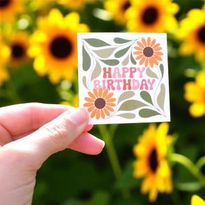 May include: A square sticker with the words "HAPPY BIRTHDAY" in pink and orange retro font. The sticker features two orange and brown flowers, green leaves, and a white background. The sticker is held in front of a background of yellow sunflowers.