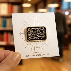 May include: A black and gold enamel pin with the text "Emotionally Attached to Fictional Characters" on a white card. The card also reads "Just One More Chapter". The pin is held in front of a blurred bookstore background.