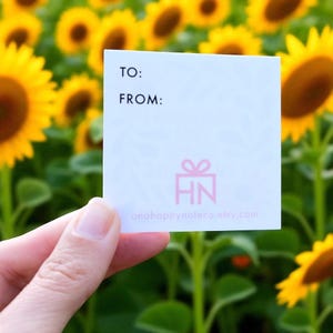 May include: A white square gift tag with the words "TO:" and "FROM:" in black. Below is a pink gift icon and the letters "HN". The website address onahappynoteco.etsy.com is printed at the bottom. The background is a field of sunflowers.