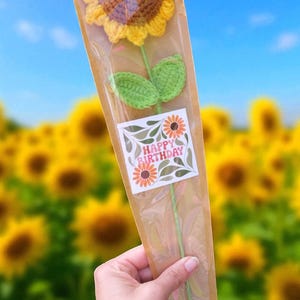 May include: A handmade crochet sunflower with yellow petals and a brown center, green leaves, and a green stem. The flower is packaged in a clear cellophane sleeve with a "Happy Birthday" sticker. The background is a field of sunflowers.