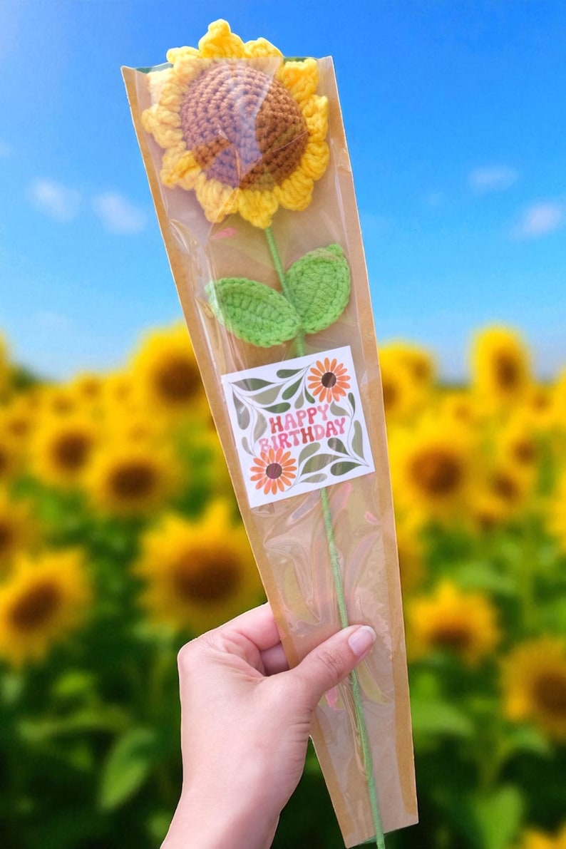 May include: A handmade crochet sunflower with yellow petals and a brown center, green leaves, and a green stem. The flower is packaged in a clear cellophane sleeve with a "Happy Birthday" sticker. The background is a field of sunflowers.