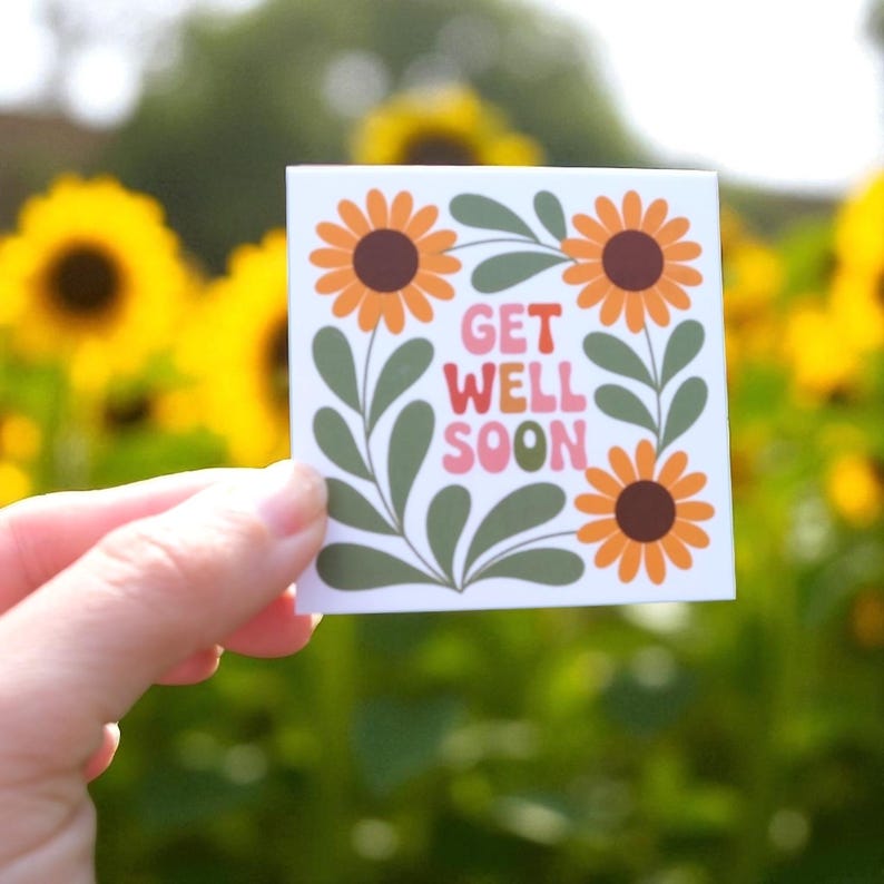 May include: A square sticker with a floral design and the words "GET WELL SOON." The sticker features orange sunflowers with brown centers, green leaves, and the text in pink and white. The background is a blurred view of sunflowers.