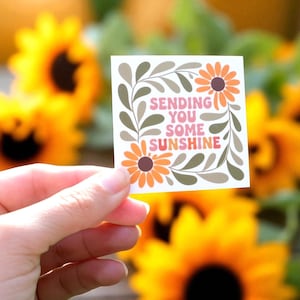 May include: A square sticker with the text "SENDING YOU SOME SUNSHINE" in pink and orange. The sticker features orange flowers and green leaves, held in front of a blurred background of yellow sunflowers.
