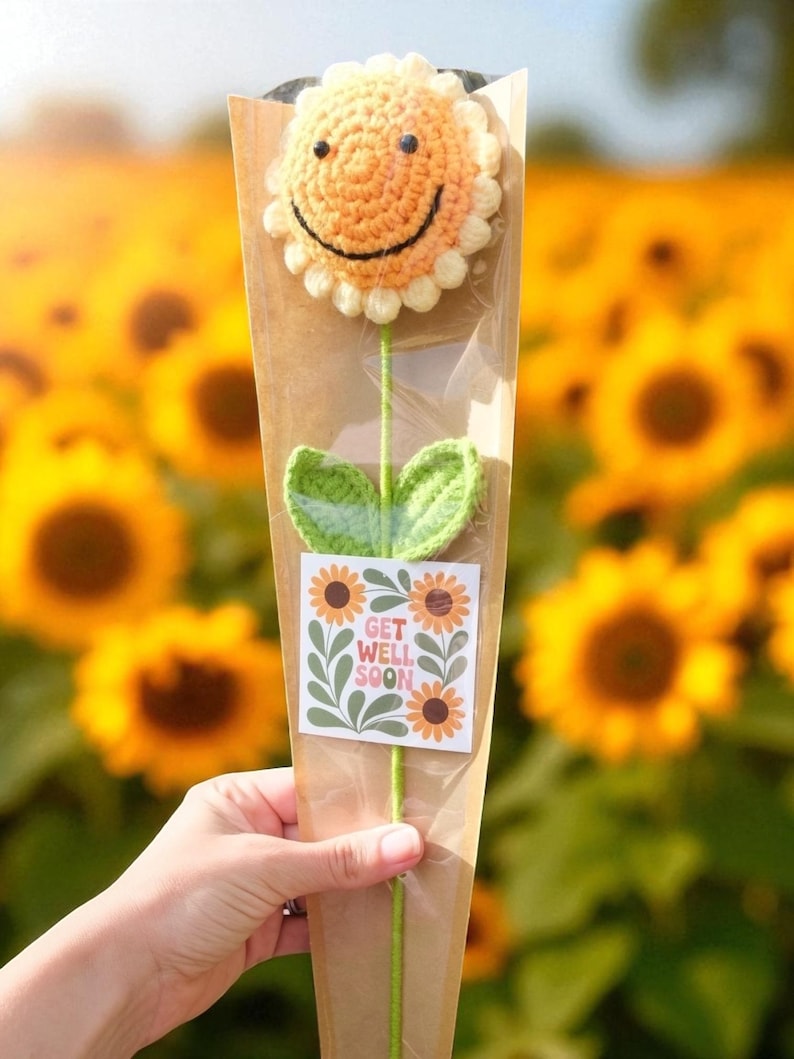 May include: A crocheted sunflower with a smiling face, green leaves, and stem, presented in a brown paper cone. A sticker with the words "Get Well Soon" and sunflower graphics is attached. The background is a field of sunflowers.