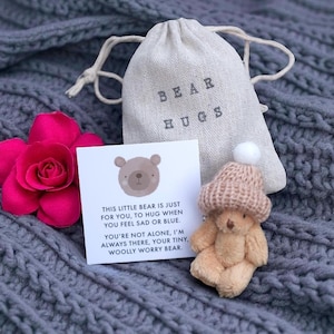 May include: A beige teddy bear wearing a knitted hat with a white pom-pom, beside a card and a drawstring bag. The card has a bear illustration and text that reads, "This little bear is just for you, to hug when you feel sad or blue."