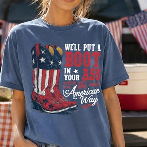 May include: Blue jean t-shirt with a graphic of a red cowboy boot with stars and stripes, and the text "We'll put a boot in your ass the American way". The shirt is worn by a person.