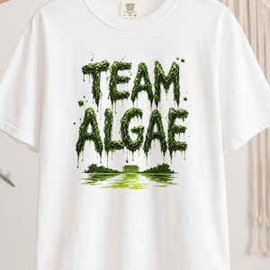 May include: A white short-sleeve t-shirt featuring a graphic print with the text TEAM ALGAE in a dripping, textured green font. Below the text is a green illustration of a building reflected in water, creating a swamp-like visual effect.