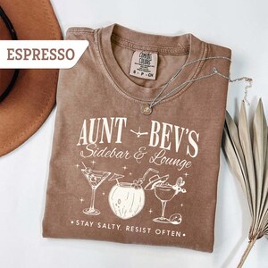 May include: Espresso-colored t-shirt with the text "Aunt Bev's Sidebar & Lounge" and cocktail illustrations. The shirt also features the phrase "Stay Salty, Resist Often." A silver necklace and a hat are also in the image.