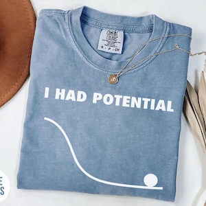 May include: A blue t-shirt with the text "I HAD POTENTIAL" in white. A white graphic of a ball rolling down a curved path is below the text. A gold necklace and dried flowers are on the shirt. The shirt is folded.