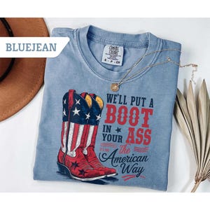 May include: Blue jean colored t-shirt with a graphic of a red and white cowboy boot with stars and stripes. The text on the shirt reads "We'll put a boot in your ass the American way."