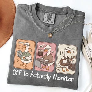 Op de afbeelding: Een grijs Comfort Colors t-shirt met een afbeelding van een gans met een bril in drie verschillende tech-gerelateerde scenario's. De tekst "Off To Actively Monitor" staat eronder. Een zilveren ketting en handschoenen zijn ook zichtbaar.