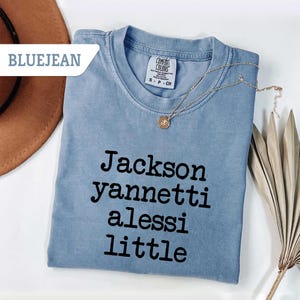 May include: A bluejean-colored t-shirt with the words "Jackson yannetti alessi little" printed in black. A gold necklace with a pendant rests on the shirt. The shirt is a Comfort Colors brand, size S-P-CH.