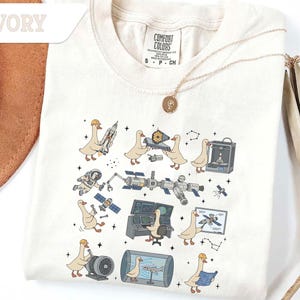 May include: Ivory-colored t-shirt featuring a whimsical design of cartoon geese interacting with space-themed elements like rockets, satellites, and a 3D printer. The shirt has a crew neck and short sleeves. The text "IVORY" is at the top left.