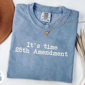 May include: A blue Comfort Colors t-shirt with the text "It's time 25th Amendment" in white. A gold necklace with a pendant rests on the shirt. A brown hat and dried flowers are also in the image.