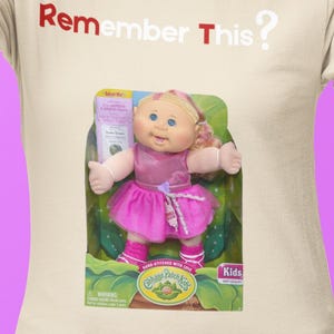 Może przedstawiać: Kremowa koszulka z napisem "Remember This?" w kolorze czerwonym i białym. Koszulka przedstawia lalkę Cabbage Patch Kids w oryginalnym opakowaniu. Lalka ma na sobie różową sukienkę i skarpetki.