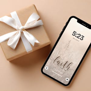 May include: A wrapped gift box with a white satin ribbon and a smartphone. The phone displays the time, date, and the words "faith over fear" on the screen. The background is a neutral beige color.