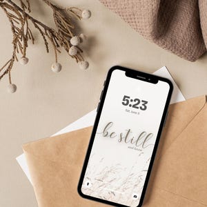 May include: A black smartphone displays a wallpaper with the words "be still and know" in a cursive font, along with the time and date. The phone rests on a brown envelope and white paper, with a beige background and decorative sprig.