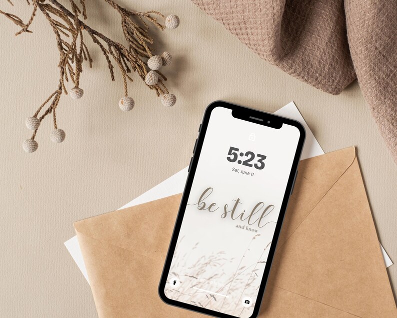May include: A black smartphone displays a wallpaper with the words "be still and know" in a cursive font, along with the time and date. The phone rests on a brown envelope and white paper, with a beige background and decorative sprig.