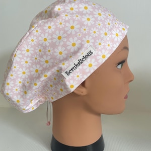 May include: A scrub cap featuring a light pink background with a repeating pattern of white daisies with yellow centres. A white fabric tag with the black text Scrubalicious is sewn onto the side. The cap includes a drawstring with a toggle.