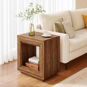 May include: A square, wooden side table with a warm brown finish. The table has an open center and a lower shelf. A small vase with greenery and a cup of coffee sit on top. The table is next to a cream-colored sofa.