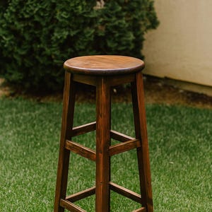 May include: A tall, wooden bar stool with a round seat and four sturdy legs. The stool is stained a rich, dark brown color, and the design includes supportive crossbars. It is set on a green lawn.