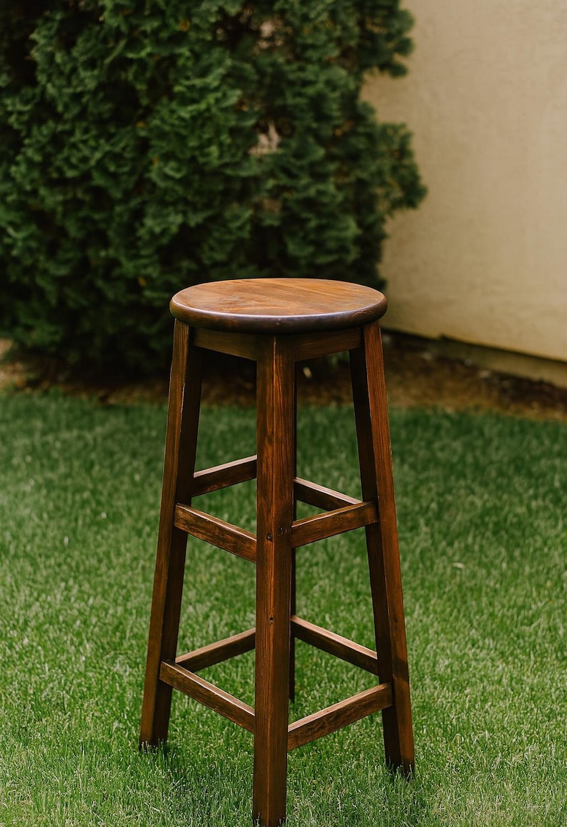 May include: A tall, wooden bar stool with a round seat and four sturdy legs. The stool is stained a rich, dark brown color, and the design includes supportive crossbars. It is set on a green lawn.