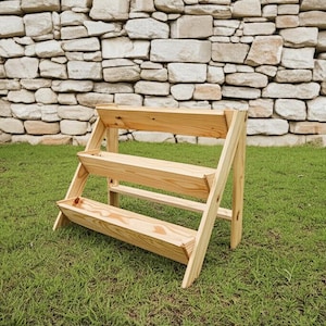 Peut inclure: Un bac à plantes en bois à trois niveaux se dresse sur l'herbe verte devant un mur de pierres. Le bac à plantes en bois naturel comporte trois bacs rectangulaires pour les plantes, soutenus par des pieds inclinés. Le design est simple et rustique.