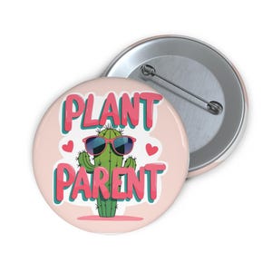 May include: A round, light pink button with the words "PLANT PARENT" in pink and teal lettering. A cartoon cactus wearing sunglasses is in the center, with two red hearts on either side. A second button is visible in the background.