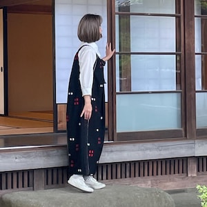 May include: A person wearing a black jumpsuit with a white shirt underneath, standing on a stone in front of a traditional Japanese building. The jumpsuit has a pattern of white and red squares. The building has wooden sliding doors and a tiled roof.